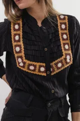 Camisa negra de tela texturizada con rayas verticales, cuello camisero y cierre frontal con botones. Presenta un diseño distintivo con un peto con tablas asimétricas y un aplique decorativo de crochet en diseño granny square en tonos mostaza, marrón y blanco.