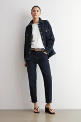 Camisa de jean azul oscuro con cierre frontal de botones metálicos a presión y dos bolsillos delanteros con solapa en el pecho. Presenta mangas largas con puños anchos doblados y cuello camisero clásico.