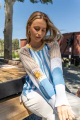 Sweater tejido a rayas verticales celestes y blancas, con cuello redondo amarillo y detalle bordado del sol de la bandera uruguaya en la manga.
