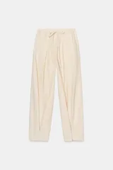 Pantalón de tiro medio color beige claro, de corte ancho y fluido, con pliegues marcados en el delantero y cintura ajustable con cordones. Presenta un detalle de pliegue con botón en el bajo.