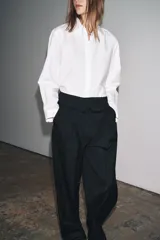 Camisa blanca de popelín de algodón 100%, con cuello solapa, manga larga terminada en puño y cierre frontal con botones ocultos por solapa. Presenta un corte holgado y ruedo curvo.