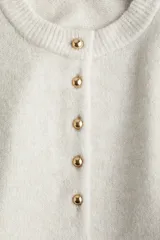 Buzo de punto suave color beige claro, con cuello redondo y abertura frontal con botones dorados. Presenta hombros caídos y puños y bajo acanalados.