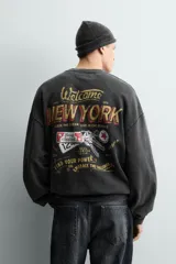 Sudadera de corte relajado, color negro con efecto lavado, cuello redondo y manga larga. Presenta un pequeño estampado frontal con texto en tonos naranja y amarillo.