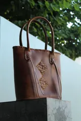 Bolso tipo matera de cuero uruguayo color marrón, con diseño artesanal de tejido trenzado en el frente y detalles de costuras decorativas en los bordes superiores.