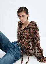 Blusa de gasa semitransparente con estampado floral en tonos rosa, verde y negro. Tiene cuello en V con lazo, cierre frontal con botones, mangas largas abullonadas con puños elásticos y corte recto.