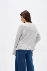 Saco de punto color gris, con cuello redondo, cierre frontal con botones y detalle de volados en la tapeta y el ruedo. Presenta mangas largas con puños acampanados.