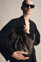 Bolso tipo saca de piel color marrón oscuro, con cuerpo fruncido y doble asa de mano rígida metálica. Incluye asa extraíble para llevar al hombro o cruzado y bolsillo interior extraíble.