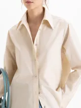 Camisa de poplin color crema, de corte clásico con cuello camisero, mangas largas y cierre frontal con botones dorados de media esfera.