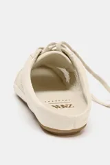 Zapato deportivo tipo mule, color crema o beige claro, con diseño destalonado. Presenta paneles de malla transpirable combinados con apliques de cuero liso y texturizado, y cordones en la parte delantera. La suela es plana de goma color caramelo.