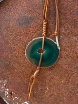 Collar con dije de piedra ágata blanca y marrón, con forma circular irregular y agujero en el centro. El dije está sujeto con un cordón fino de cuero marrón.