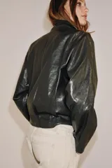 Chaqueta de corte amplio estilo biker en tejido efecto piel de color verde oscuro, con cuello alto envolvente, cierre frontal asimétrico de cremallera metálica, bolsillos ocultos en costura y forro interior.