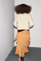 Trench corta color beige con cuello solapa, manga larga con trabilla y botón en los puños, bolsillos de vivo en el delantero y cierre frontal con botones oscuros.