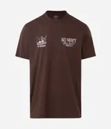 Remera de algodón color marrón con cuello redondo y mangas cortas. Presenta un diseño gráfico de estilo barroco con una figura alada y la inscripción 'Humble' en la espalda.