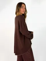 Sweater de punto gris con cuello redondo, mangas largas y diseño asimétrico, siendo más corto en la parte delantera y más largo en la trasera, con detalles de terminación en contraste.