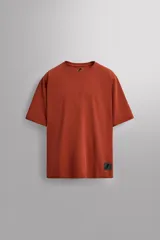 Camiseta de ajuste relajado (relaxed fit) color terracota, confeccionada en tejido técnico. Presenta cuello redondo, mangas ranglán cortas y un parche negro con logo en el bajo.
