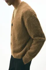 Cárdigan de punto marrón con textura peluda, cuello en V pronunciado y cierre frontal de botones. Presenta remates acanalados en puños y bajo, con un corte holgado.