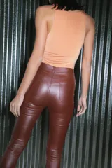 Pantalón de tiro alto confeccionado en bengalina engomada con acabado brillante. Presenta un diseño ajustado tipo chupín con cierre frontal, botones y remaches metálicos.