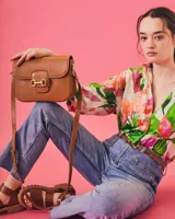 Cartera negra de cuero charolado con solapa y cierre de broche. Presenta un detalle metálico dorado en forma de freno de caballo en la parte frontal y una correa ajustable.