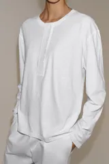 Camiseta blanca de manga larga y corte regular fit, confeccionada en mezcla de algodón y lino. Presenta cuello panadero con cierre frontal de botones y bajo recto.