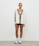 Cardigan deportivo de corte oversized, color blanco con detalles en marrón oscuro. Presenta escote en V, cierre frontal con botones a presión, mangas largas con franjas laterales y puños acanalados. Incluye un parche circular con letras en el pecho.