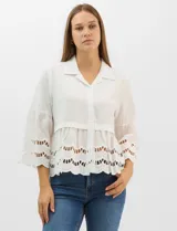 Camisa blanca de corte recto con cuello camisero y cierre frontal de botones. Presenta mangas tres cuartos acampanadas con detalles de bordado suizo calado en los puños y en el ruedo inferior.