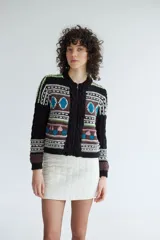 Campera de punto jacquard con lana merino, diseño con cierre frontal, cuello redondo y detalles de trenzas en los laterales. Presenta un patrón geométrico multicolor con tonos marrones, negros, azules y verdes.
