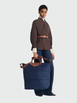 Bolso de viaje grande de la línea Le Pliage Original, color azul marino. Presenta un diseño estructurado con asas cortas de cuero marrón, solapa con cierre de botón a presión y correa de hombro ajustable y extraíble. El material principal es lona resistente con detalles en cuero vacuno.