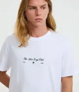 Remera masculina color blanco, modelo Comfort, de manga corta y cuello redondo. Presenta una pequeña estampa frontal con texto y una estampa grande de palmeras en la espalda.