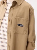 Camisa de gabardina color verde oliva, con cuello clásico, cierre frontal de botones y dos bolsillos frontales con parche. Incluye un pequeño parche con el logo de la marca en el bolsillo izquierdo.