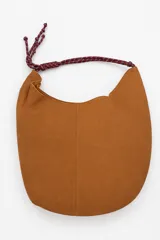Cartera tipo hobo de gamuza color marrón, con costura central vertical y correa de cuerda trenzada en tonos borró y gris con nudos decorativos en los extremos.
