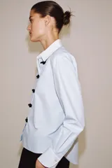 Camisa de algodón con cuello solapa y puños en color blanco a contraste. Presenta cierre frontal con alamares negros y bajo asimétrico redondeado.