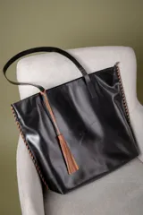 Bolso tipo shopper confeccionado en cuero vacuno negro, con detalles de costura artesanal en los bordes en color suela. Incluye un cierre superior y un adorno de borla de flecos de cuero en color suela colgando de una de las asas.