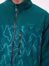 Campera reversible con cuello alto y cierre frontal. Un lado presenta tejido de polar con estampado geométrico en tonos verdes y bolsillo con cierre en el pecho, mientras que el otro lado es de nylon. Cuenta con puños elásticos y bolsillos laterales.