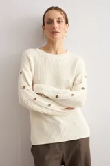 Sweater de punto color blanco natural, con cuello redondo y mangas largas adornadas con botones dorados a lo largo de las costuras exteriores.