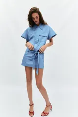 Vestido corto de estilo camisero color azul, con cuello solapa, manga corta y cierre frontal con botones. Presenta un bolsillo de plastrón con solapa en el pecho y un cinturón ajustable del mismo tejido en la cintura.