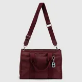 Bolso tipo tote de la marca Bubba, color verde petróleo, con doble asa de mano y correa larga ajustable y extraíble con logo grabado. Incluye cierre principal, bolsillo interior y un dije colgante con la letra B en color rosado.
