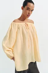 Blusa de lino color amarillo pastel, con escote recto ajustable mediante lazadas, hombros descubiertos y mangas largas abullonadas con puños elásticos.