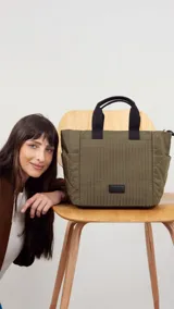 Cartera tipo tote de diseño capitoneado en color marrón claro, con doble asa de mano negra y bolsillos laterales. Presenta un diseño acolchado con líneas verticales en el frente y rombos en los laterales, e incluye una etiqueta decorativa con logo.