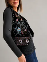 Chaleco de estilo boho chic confeccionado en tela aterciopelada negra, con bordados florales multicolor y detalles de tachas metálicas a lo largo de los bordes frontales.