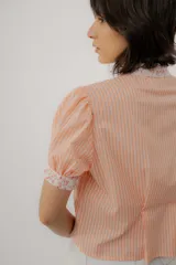 Blusa celeste a rayas blancas, de manga corta abullonada. Cuenta con cuello mao y tapeta de botones con detalles de estampado floreado en tono rosado, presentes también en el cuello y los puños.