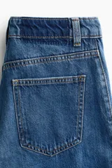 Jean de tiro alto y corte acampanado, confeccionado en denim de algodón. Presenta diseño de cinco bolsillos, cierre frontal con botón y costuras decorativas laterales.
