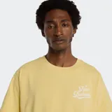 Remera de algodón color amarillo pálido con cuello redondo y manga corta. Presenta un pequeño estampado en el pecho con el logo de New Balance en tipografía cursiva blanca.