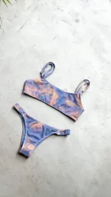 Conjunto de bikini con corpiño estilo bandó alto y breteles regulables, con lazo para atar en la espalda. La bombacha es vedetina con frunce elástico en los costados. Presenta un estampado tipo tie-dye o marmolado en tonos azules y naranjas/terracota.