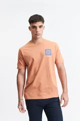 Remera de manga corta color salmón, con escote redondo y un pequeño estampado frontal de una flor azul sobre un fondo texturizado, con la inscripción "MISTRAL ® 1976 INTERNATIONAL".