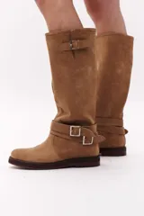 Bota de caña alta color marrón chocolate, confeccionada en gamuza. Presenta dos correas desmontables con hebillas metálicas plateadas.