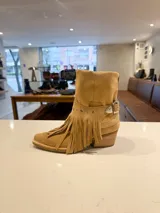 Bota de caña alta estilo western confeccionada en cuero vacuno color suela. Presenta un diseño versátil con estribo desmontable y flecos decorativos en la zona del tobillo, taco de madera y punta fina.
