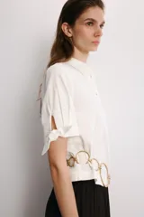 Camisa de manga corta confeccionada en tejido de ramio color blanco. Posee cuello solapa, cierre frontal con botones y detalle de lazos en el acabado de las mangas.