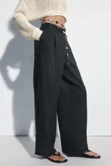 Pantalón de lino negro de tiro medio, con pernera recta, pinzas delanteras y detalle de puntilla en la cintura. Cuenta con bolsillos frontales y cierre con cremallera y botón.