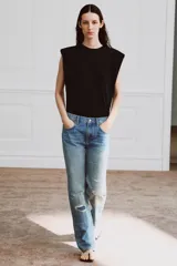 Jeans de tiro medio con corte recto y largo completo. Presentan un diseño de cinco bolsillos, cierre frontal con cremallera y botón, y un acabado con efecto lavado. Incluyen detalles de roturas y parches decorativos en la parte delantera.