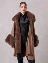 Ruana de tejido color tostado con cuello, bordes frontales y puños amplios con apliques de piel sintética al tono. Presenta una abertura central con cierre de botón tipo trenca.